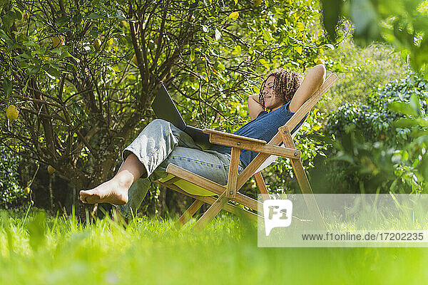 Lächelnde mittelgroße erwachsene Frau mit Händen hinter dem Rücken  die auf einen Laptop schaut  während sie auf einem Stuhl im Garten sitzt
