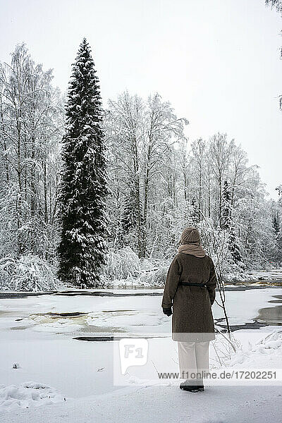 Junge Frau in warmer Kleidung steht an einem zugefrorenen Fluss im Wald