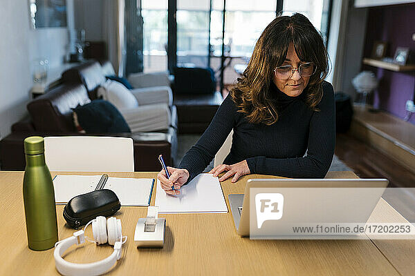 Reife Frau schreibt in Papier  während sie zu Hause am Laptop sitzt