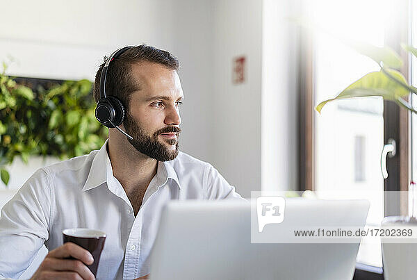 Nachdenklicher Geschäftsmann mit Kopfhörern  der im Büro am Laptop sitzt