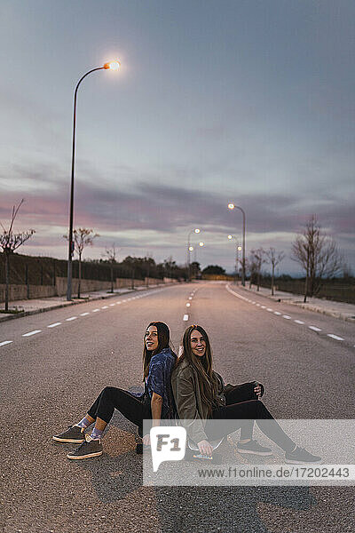 Lächelnde Freundinnen sitzen in der Abenddämmerung Rücken an Rücken auf einem Skateboard