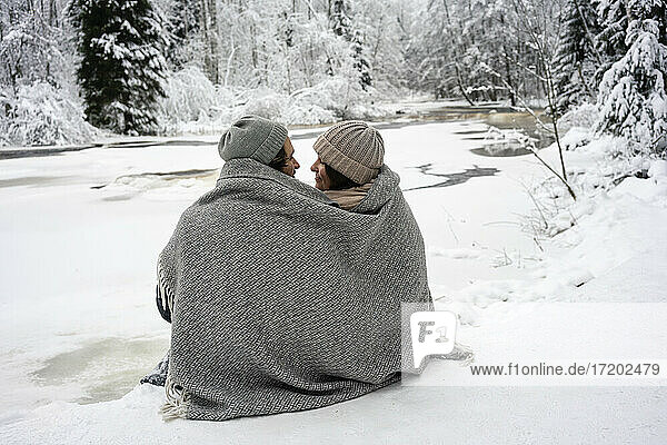 Junges Paar  in eine Decke gehüllt  sieht sich gegenseitig an  während es an einem zugefrorenen Fluss im Wald sitzt