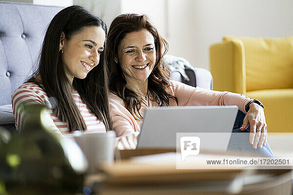 Lächelnde Mutter und Tochter schauen gemeinsam auf den Laptop  während sie zu Hause im Wohnzimmer sitzen