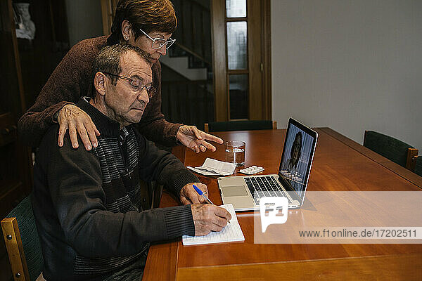 Älterer Mann mit Sozialarbeiter beim Schreiben während einer Videokonsultation mit einer Ärztin zu Hause