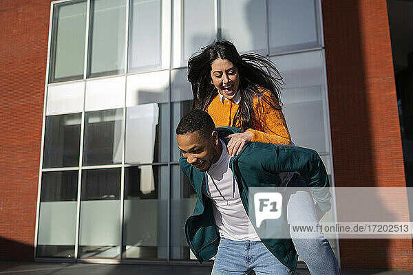 Man giving piggyback to cheerful girlfriend against modern building