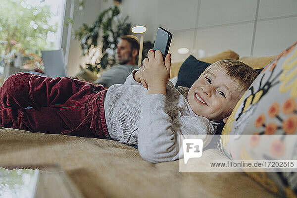 Lächelnder Junge  der ein Mobiltelefon benutzt  während er zu Hause auf dem Sofa liegt