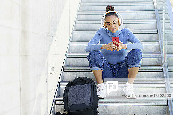 Young woman with backpack and headphones using mobile phone while sitting on steps