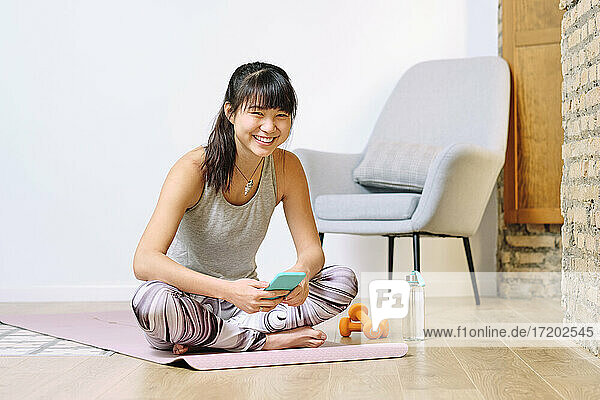 Lächelnde Frau  die ein Smartphone benutzt  während sie zu Hause im Schneidersitz sitzt