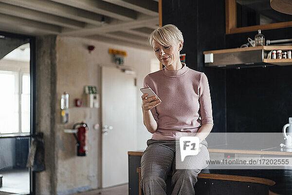 Lächelnde Geschäftsfrau mit Kaffeetasse  die ein Mobiltelefon benutzt  während sie zu Hause auf dem Küchentisch sitzt