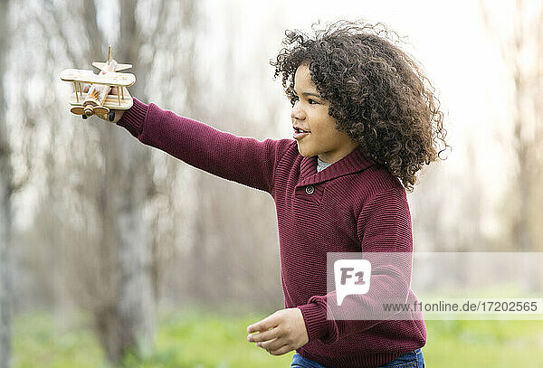 Lockenköpfiger Junge spielt mit einem Holzspielzeug-Flugzeug in der Natur