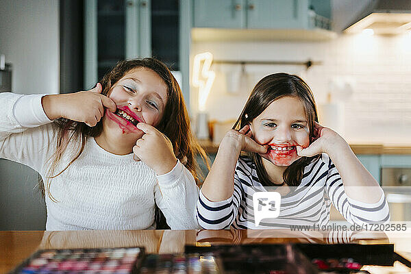 Playful girls with makeup on face teasing while sitting at dining table in domestic kitchen