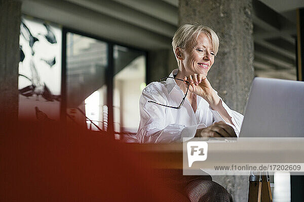 Smiling freelance worker holding eyeglasses while using laptop at desk in home office