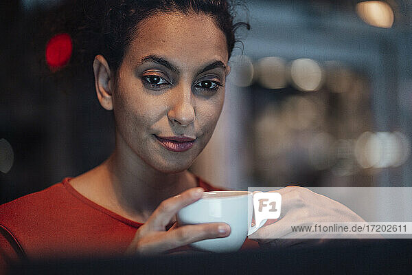 Junge Frau trinkt Kaffee  während sie in einem Café sitzt und auf ihren Laptop schaut