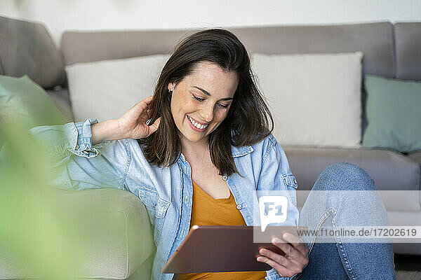 Smiling woman with hand in hair using digital tablet while sitting at sofa in living room