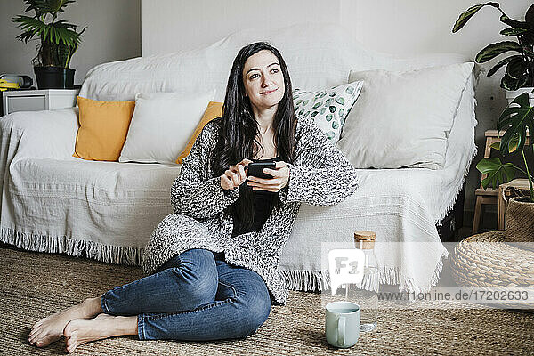 Lächelnde Frau schaut weg  während sie ihr Smartphone auf dem Sofa im Wohnzimmer benutzt