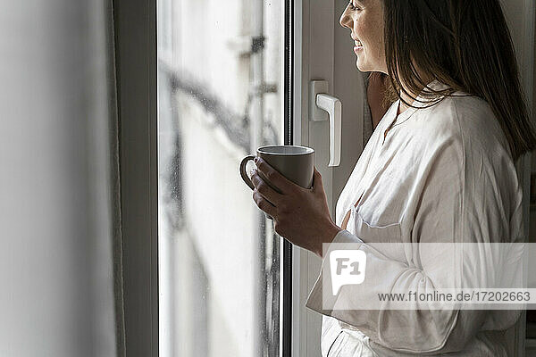Glückliche junge Frau mit Kaffeetasse  die durch das Fenster ihres Hauses schaut