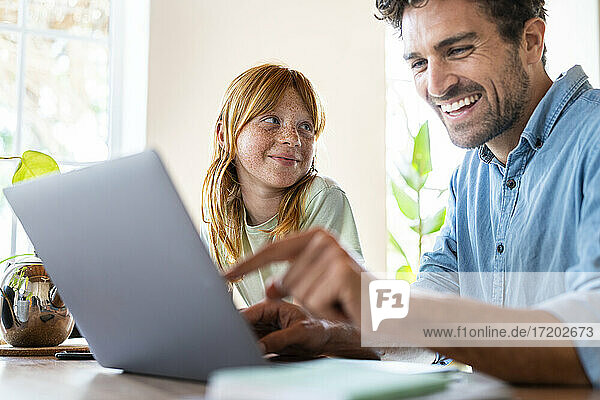 Smiling redhead girl looking at father working on laptop in living room