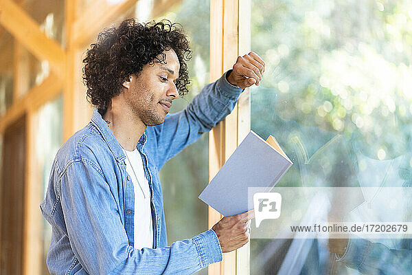 Mann liest ein Buch und lehnt sich an ein Fenster im Vorgarten