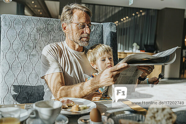 Älterer Mann mit Sohn liest Zeitung  während er am Tisch im Hotelzimmer sitzt