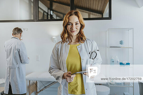 Smiling female doctor holding digital tablet while standing with coworker in background at clinic
