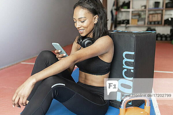 Lächelnde junge Frau  die sich an eine Box lehnt und ein Mobiltelefon im Fitnessstudio benutzt