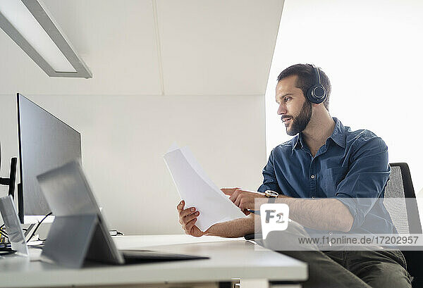 Male professional with headphones checking paper while using computer at office