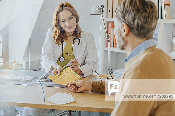 Allgemeinmediziner  der einem Patienten im Büro sitzend einen Impfpass überreicht