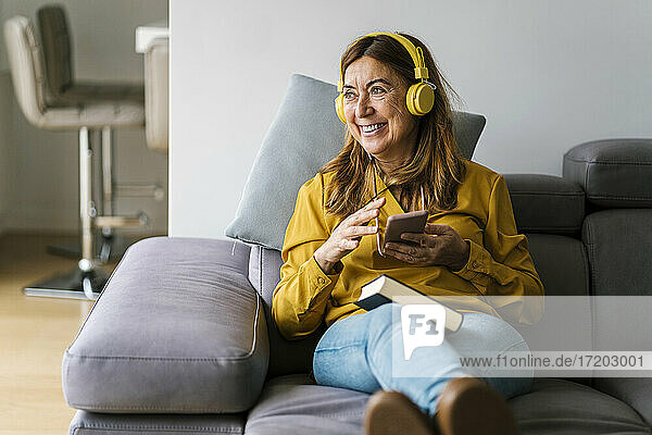 Lächelnde reife Frau  die wegschaut  während sie zu Hause über Kopfhörer Musik hört