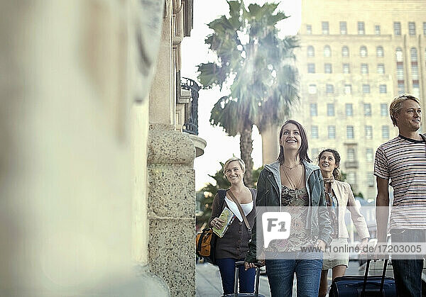 Lächelnde männliche und weibliche Freunde mit Koffern  die auf dem Gehweg einer Straße in der Stadt spazieren gehen