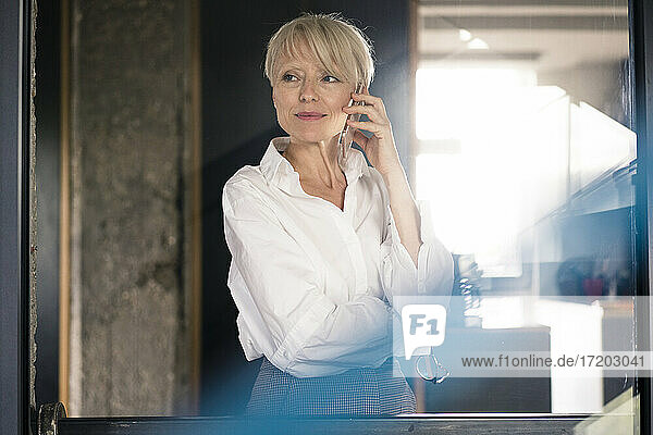 Weibliche Unternehmerin  die vor einer Glaswand im Büro zu Hause mit einem Mobiltelefon spricht