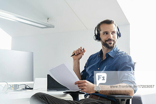 Lächelnder Geschäftsmann mit Kopfhörern und Papier im Büro sitzend