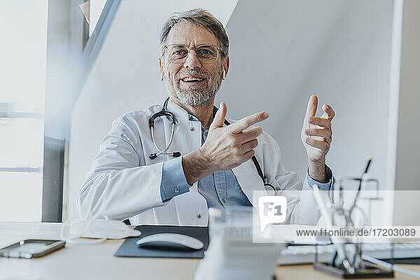 Lächelnder männlicher Arzt gestikuliert beim Sitzen in der Arztpraxis