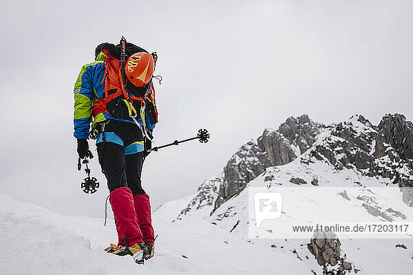 Mature man carrying backpack while walking on snowcapped mountain with crampon and hiking poles