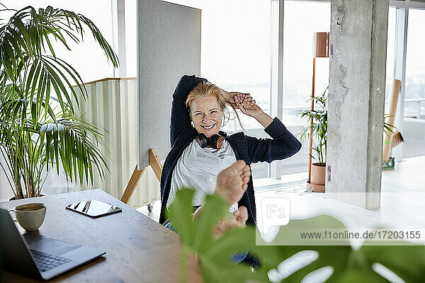 Lächelnde Unternehmerin mit Händen hinter dem Kopf  die sich auf einem Stuhl am Schreibtisch im Heimbüro entspannt
