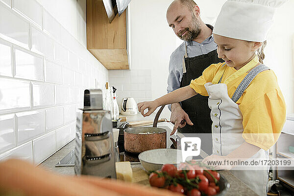 Smiling daughter wearing apron and chef's hat cooking food while standing by father in kitchen