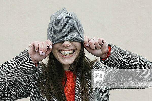 Smiling woman covering eyes through knit hat against wall