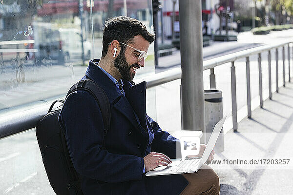 Männlicher Freiberufler arbeitet am Laptop  während er auf einer Bank am Bahnhof sitzt