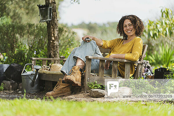 Smiling woman holding scissor while sitting on wooden bench in garden