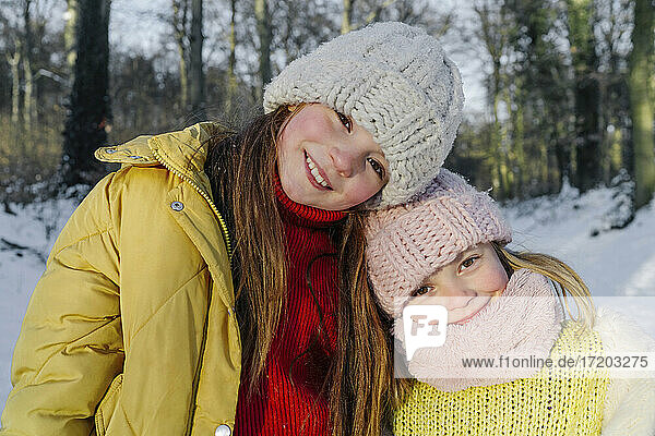Lächelnde Schwestern in warmer Kleidung  die zusammen gegen Bäume im Schnee stehen