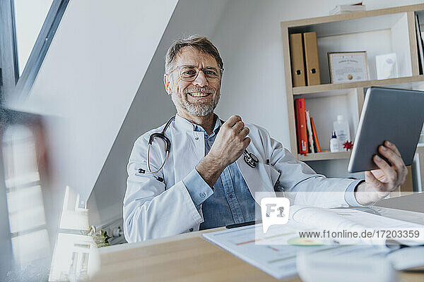 Lächelnder männlicher Arzt mit digitalem Tablet  der am Schreibtisch in einer Arztpraxis sitzt