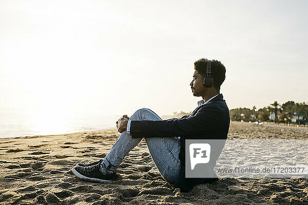Nachdenklicher Mann  der seine Knie umarmt  während er am Strand gegen den Himmel sitzt