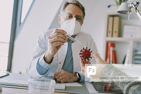 Experte mit Gesichtsschutz  der einem Patienten das Coronavirus an einem künstlichen Modell erklärt  während er im Büro sitzt