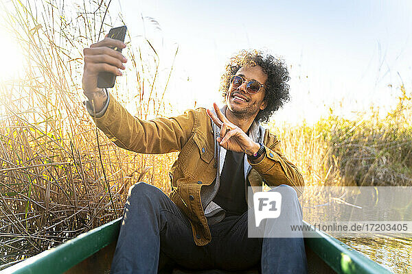 Junger Mann gestikuliert Friedenszeichen  während er ein Selfie mit seinem Handy macht  während er im Kanu auf dem Fluss sitzt