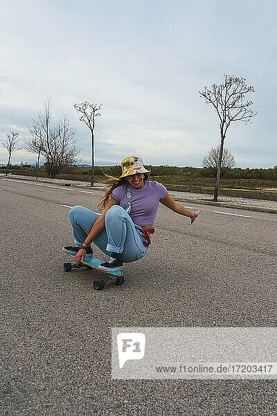 Lächelnde junge Frau hockt auf dem Skateboard gegen den Himmel