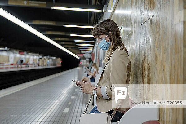 Woman with using mobile phone while sitting at subway platform during COVID-19
