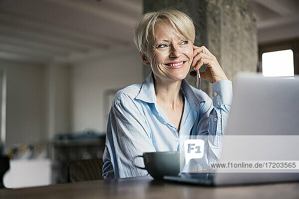 Lächelnde Geschäftsfrau mit Laptop und Kaffeetasse  die wegschaut  während sie am Schreibtisch im Heimbüro mit dem Handy telefoniert