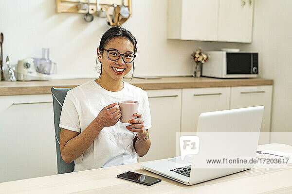 Lächelnde junge Frau sitzt mit Kaffeetasse und Laptop zu Hause