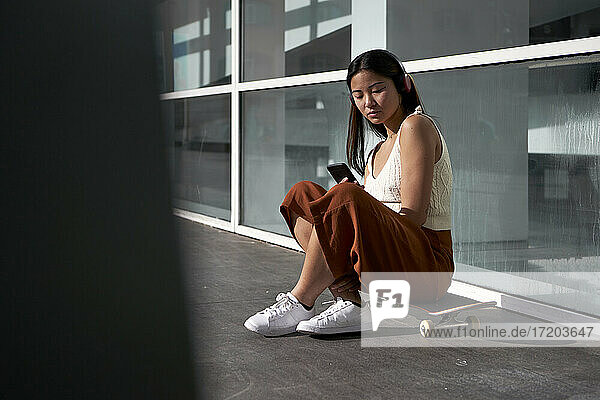 Asian woman using smart phone while sitting on skateboard during sunny day