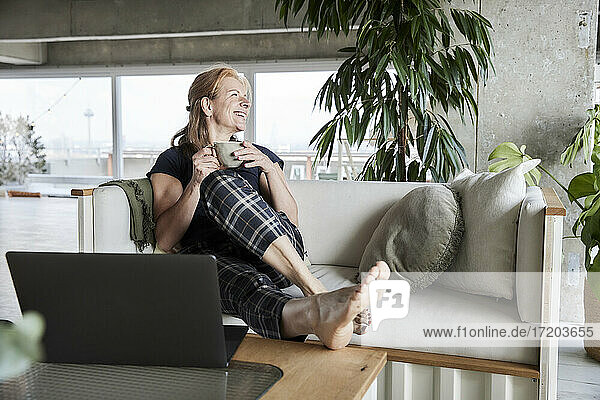 Smiling woman with laptop holding coffee cup while sitting on sofa at home