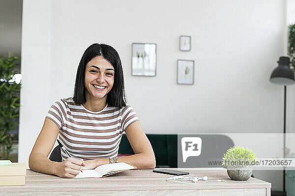 Cheerful woman with notebook and pen sitting at table in living room